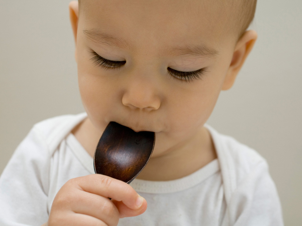 boy in white with wooden spoon to his mouth