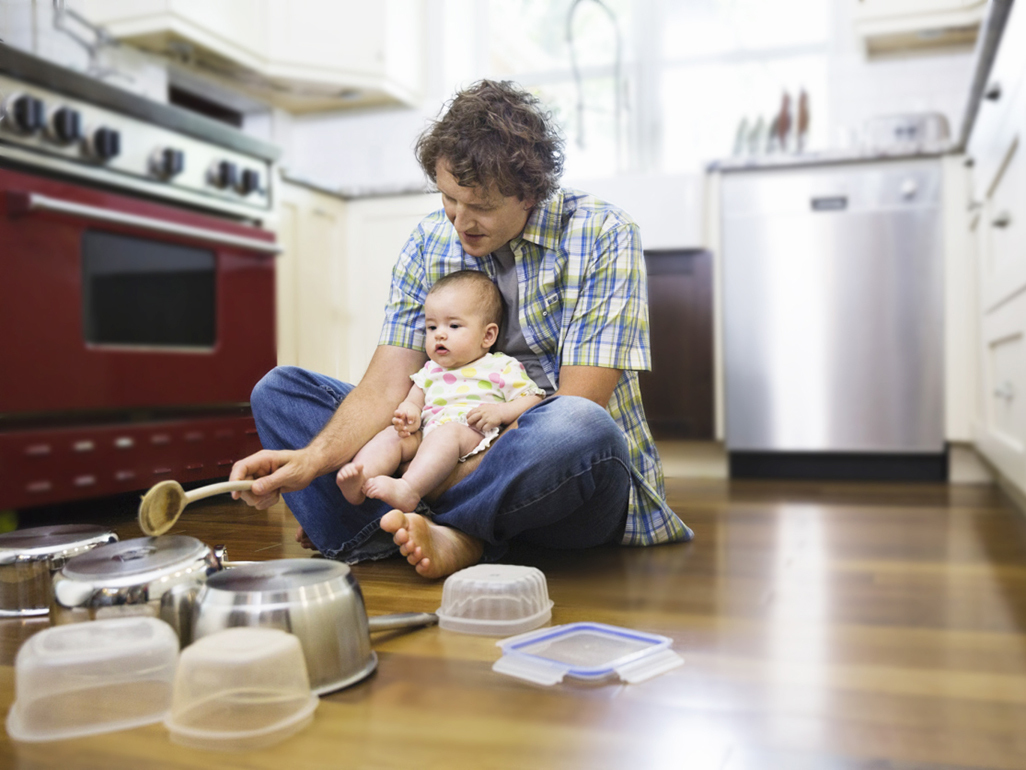 Baby sits on dads lap on the kitchen floor as they play with pots and pans