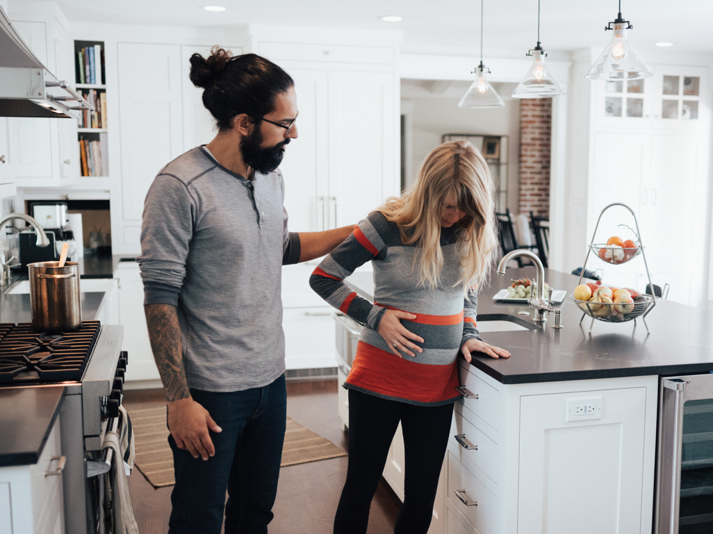Pregnant woman looking uncomfortable and feeling her bump, with partner in kitchen