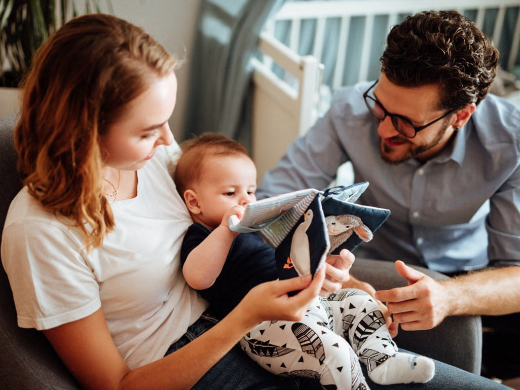 Parents smile at baby as she plays with a soft book