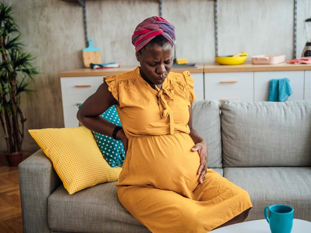 Heavily pregnant woman sitting on sofa at home, holding her bump and breathing out through pursed lips